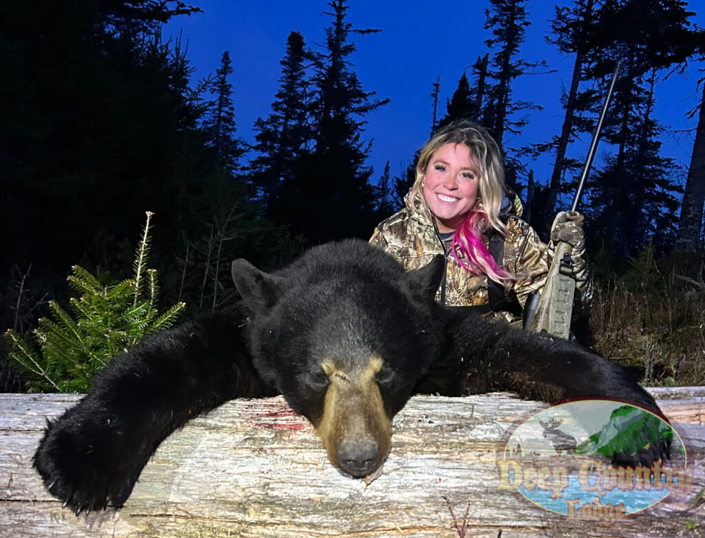 Hunter in camouflage poses with a dead black bear draped over a log in a forest at dusk, holding a rifle and smiling.