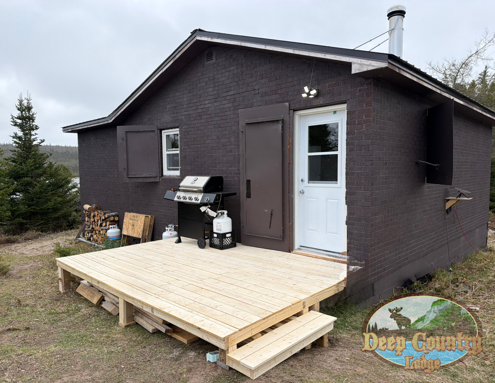 Small dark brick cabin with a wooden deck, white door, brown shutters, grill, and stacked firewood outside.