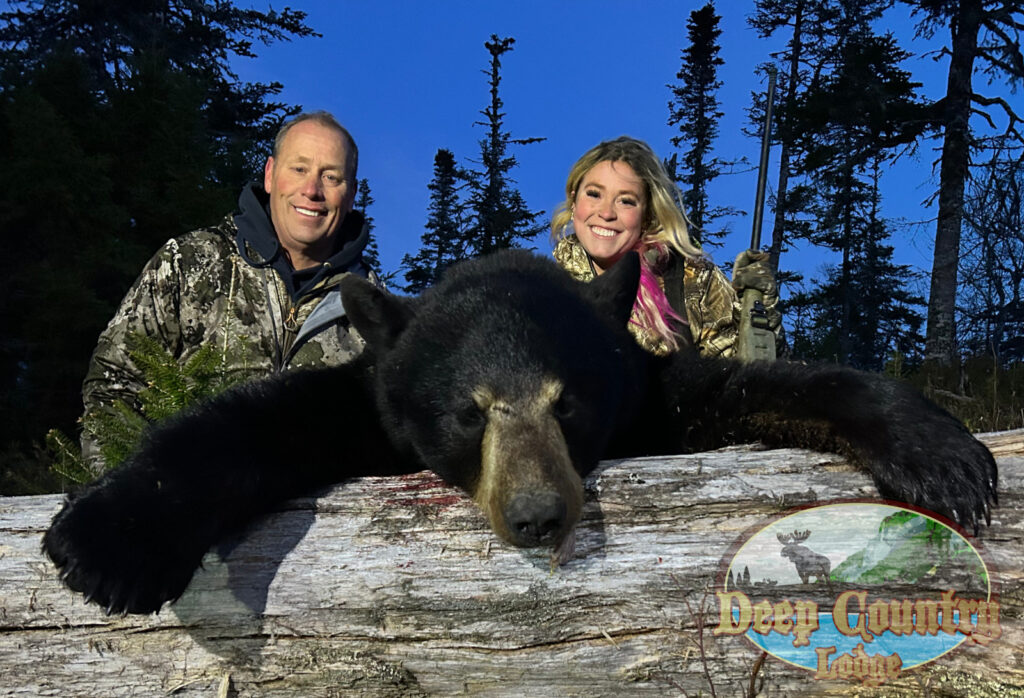 Two hunters in camouflage stand behind a large black moose carcass draped over a log in a forest, with a Deer Country Lodge logo in the bottom right.
